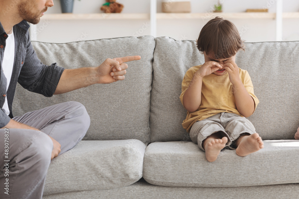 Child abuse concept. Unhappy little boy sitting on sofa and crying, unrecognizable angry father scolding at upset kid