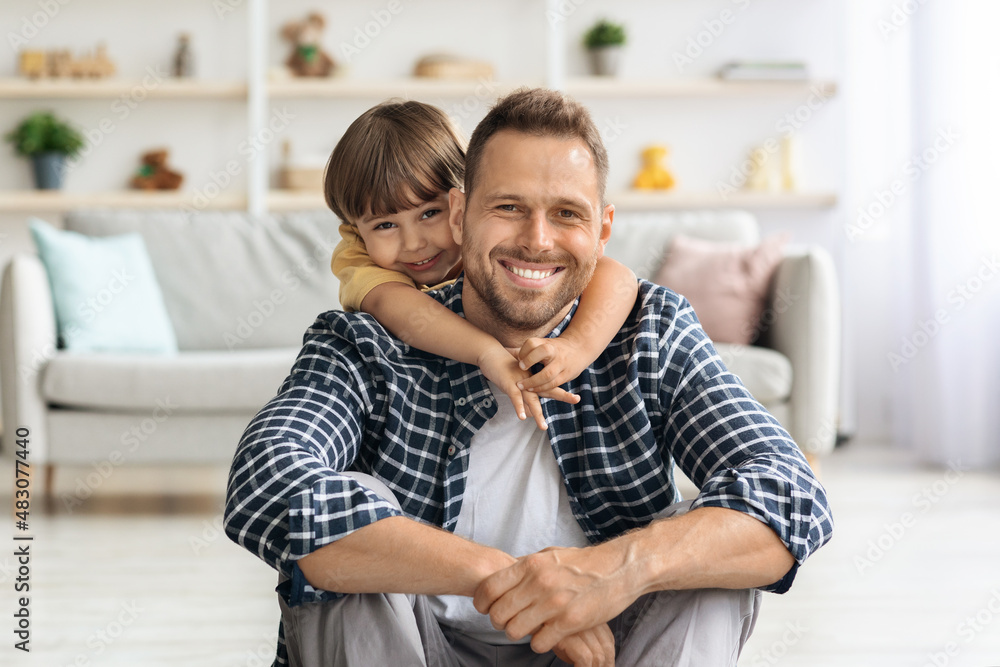 Fototapeta premium Young happy father smiling to camera, enjoying hugs of his cute little son, sitting on floor at home interior