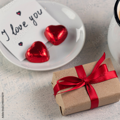 Square photo with gift with a red ribbon, a mug of coffee, heart-shaped chocolates and a note with the word I love you. Valentine's day breakfast. Valentine's Day concept. Soft focus photo