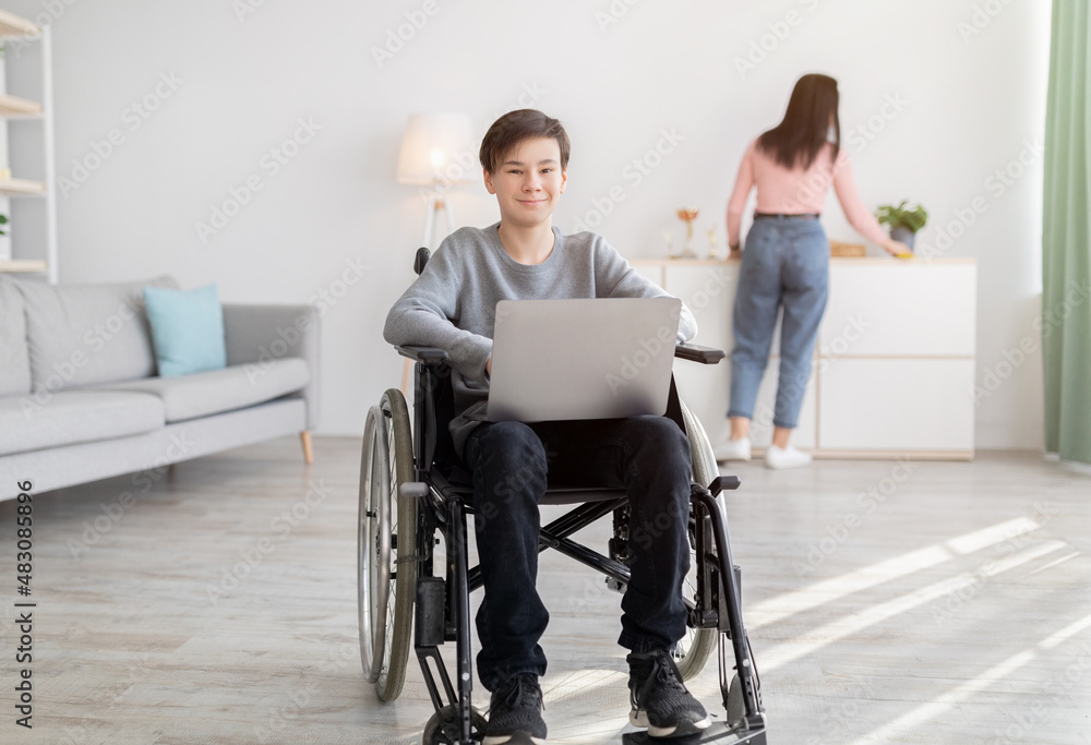Cheery disabled teen boy in wheelchair with laptop smiling at camera indoors, his mother making cleanup on background Cheery disabled teen boy in wheelchair with laptop smiling at camera indoors, his mother making cleanup on background