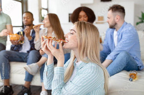 Pretty young blonde lady eating pizza on home party with her multiethnic friends, free space