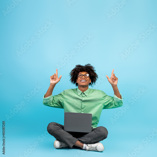 Check this. Black teen guy sitting with laptop and pointing up at free space above head over blue background, crop © Prostock-studio