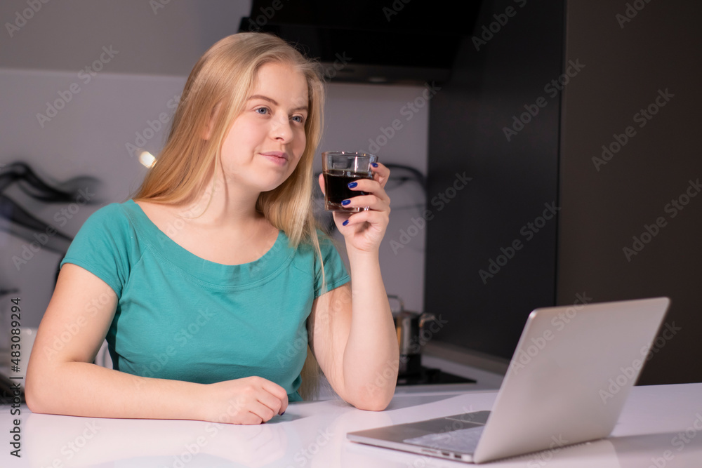pretty blonde girl in green t-shirt working or studying near laptop in grey room holding a glass with brown liquid drink. coffee tea alcohol whiskey juice pretty blonde girl in green t-shirt working or studying near laptop in grey room holding a glass with brown liquid drink. coffee tea alcohol whiskey juice