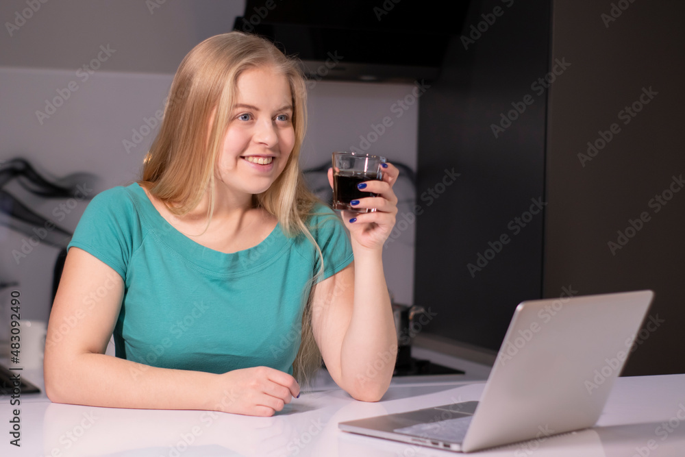 pretty blonde girl in green t-shirt working or studying near laptop in grey room holding a glass with brown liquid drink. coffee tea alcohol whiskey juice pretty blonde girl in green t-shirt working or studying near laptop in grey room holding a glass with brown liquid drink. coffee tea alcohol whiskey juice
