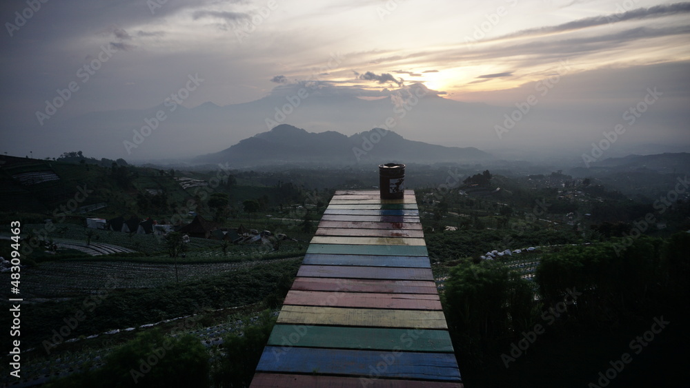Colorful bridge with sunrise sky in cloudy weather on the background. Seen mountains on the background with covered by cloud. The location named Mangli Sky View on the slope of Mount Sumbing.