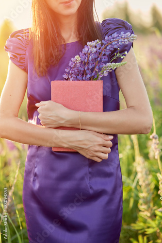 Woman in summer field with bouquet of wildflowers - lupines, hat and pink notebook. Closeness to nature, self-discovery concept. Relax and welll-being concept. Sunny day, good mood concept
