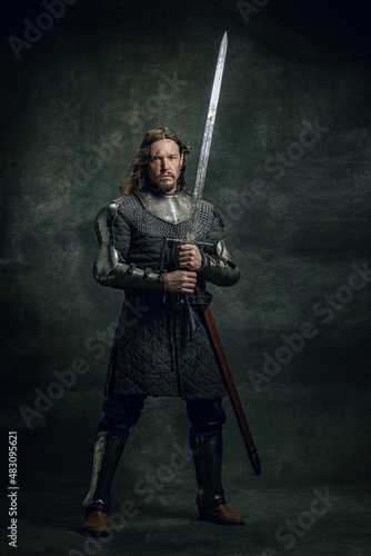 Quadro su tela Vintage style portrait of brutal seriuos man, medieval warrior or knight with dirty wounded face holding big sword isolated over dark background