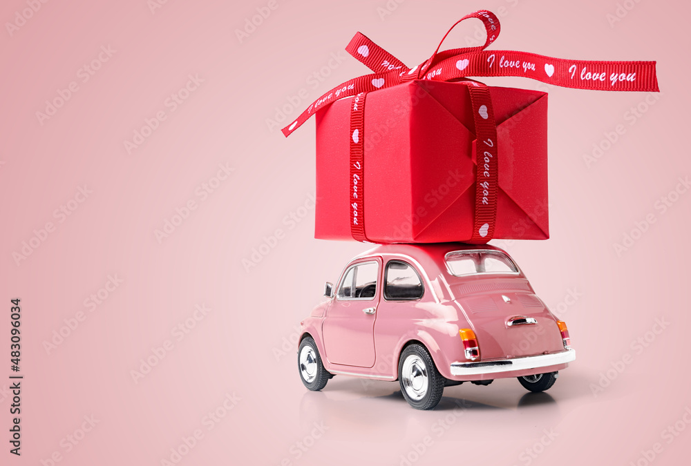 Pink retro toy car delivering red gift box with ribbon on pink background. Copy space. Pink retro toy car delivering red gift box with ribbon on pink background. Copy space.