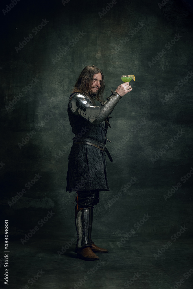 Portrait of medieval warrior or knight with dirty wounded face holding cocktail glass isolated over dark background. Comparison of eras, history Portrait of medieval warrior or knight with dirty wounded face holding cocktail glass isolated over dark background. Comparison of eras, history