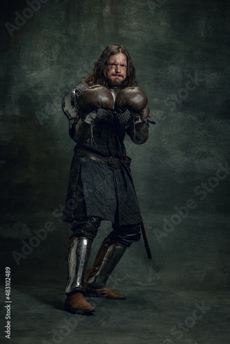 Portrait of medieval warrior or knight with dirty wounded face boxing gloves isolated over dark background. Comparison of eras, history