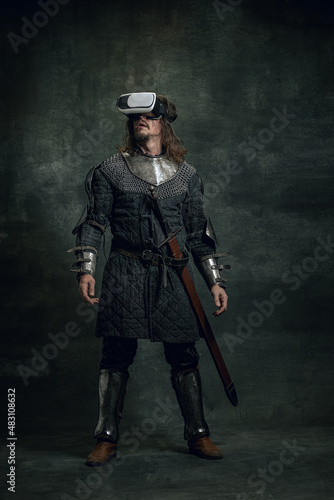Portrait of man, medieval warrior, knight with dirty wounded face in VR headset isolated over dark vintage background. Comparison of eras, history © master1305