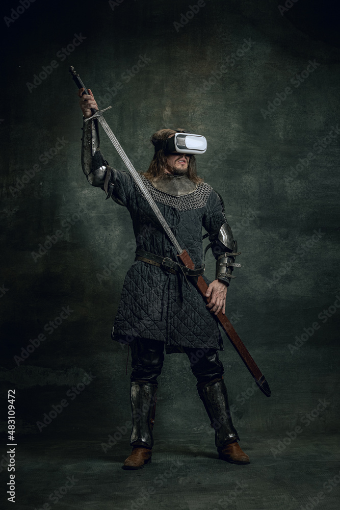 Portrait of man, medieval warrior, knight with dirty wounded face in VR headset isolated over dark vintage background. Comparison of eras, history Portrait of man, medieval warrior, knight with dirty wounded face in VR headset isolated over dark vintage background. Comparison of eras, history