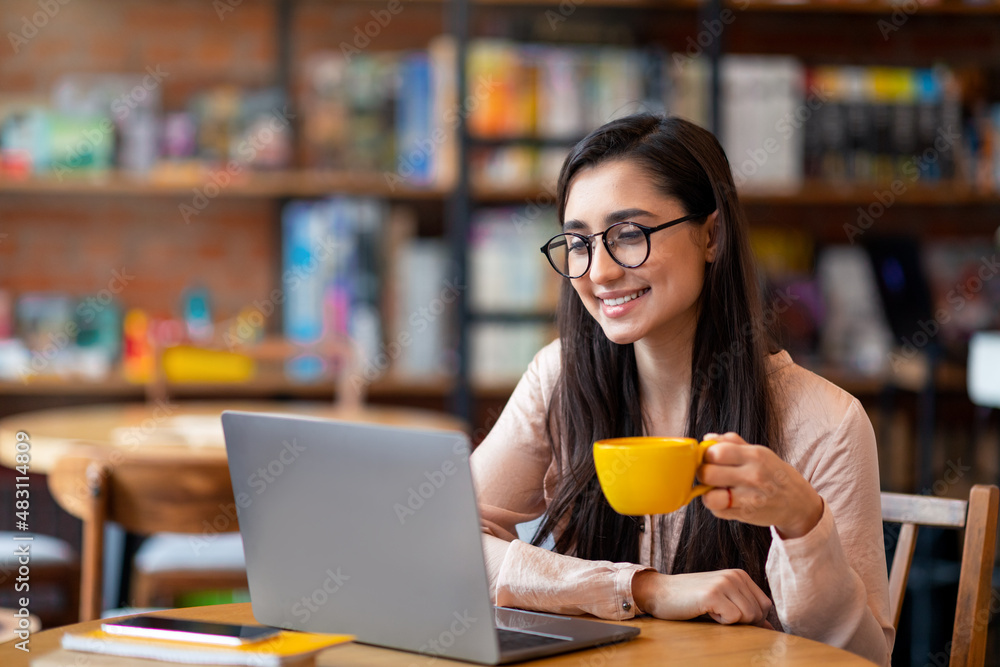 Happy latin woman working online at cafeteria, using laptop computer and drinking coffee, copy space Happy latin woman working online at cafeteria, using laptop computer and drinking coffee, copy space