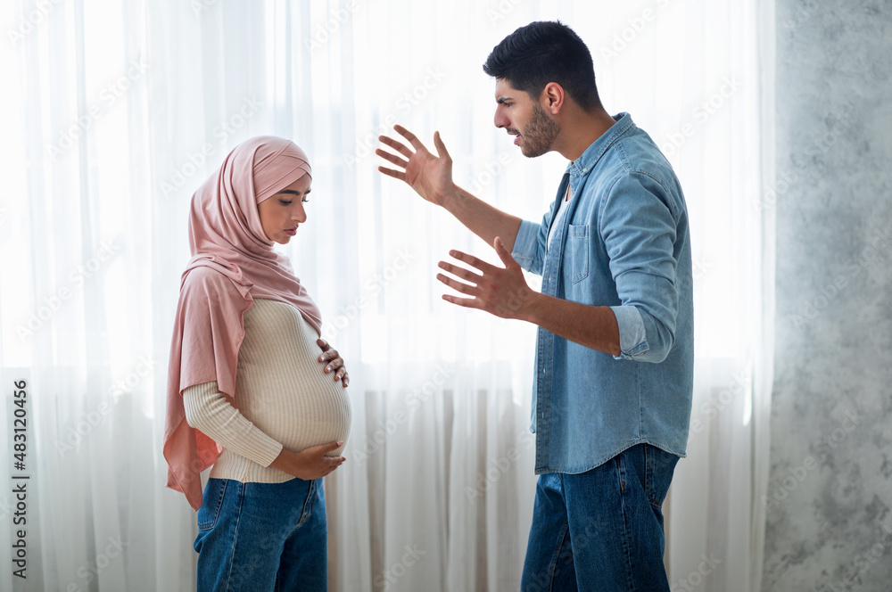 Angry arab man shouting at his pregnant muslim wife, standing near windiw at home, side view - obrazy, fototapety, plakaty 