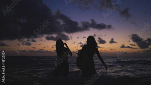 Silhouette of two young women walks up from the sea to the beach after surfboarding in the twilight time. Sunset on cloudy sky day. © suwanb