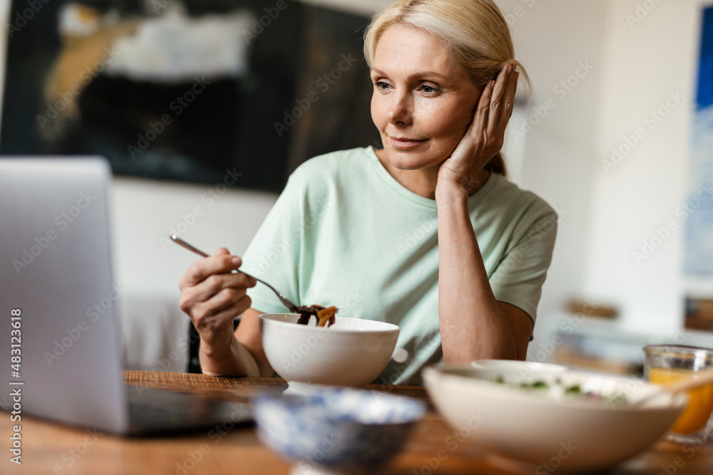 Blonde mature woman using laptop while having lunch at home