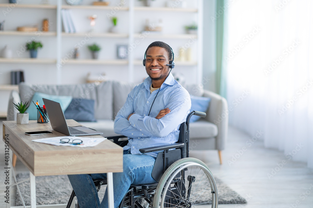 Positive paraplegic black man in wheelchair wearing headset, posing in front of laptop computer at home Positive paraplegic black man in wheelchair wearing headset, posing in front of laptop computer at home