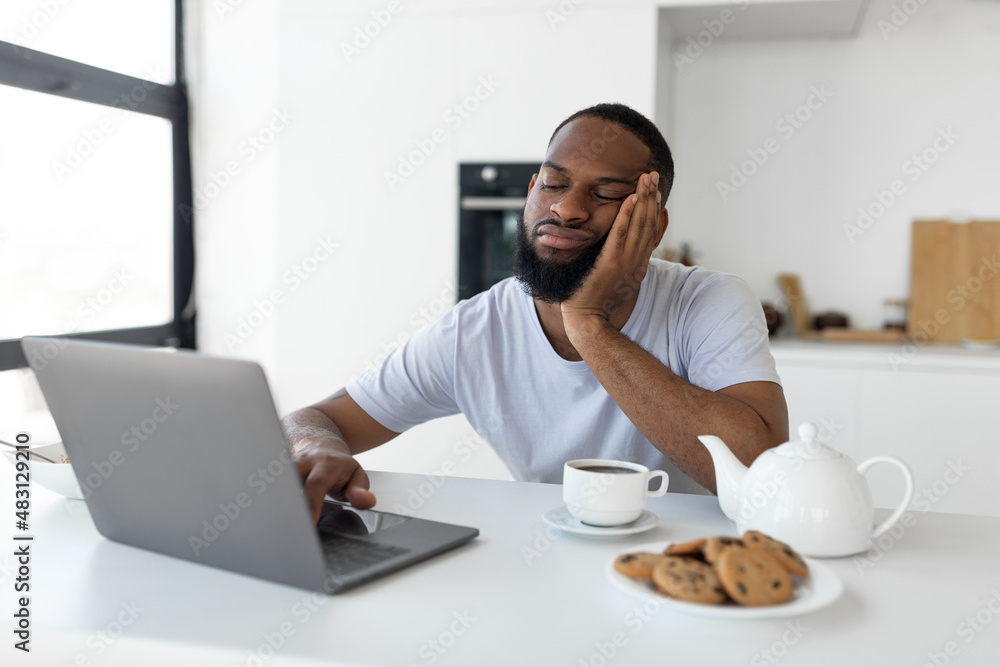 Tired black man using laptop sitting at kitchen table