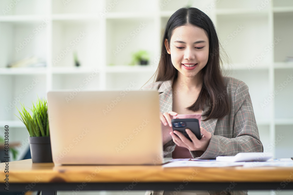 Asian woman smiling and looking at mobile phone in hand while working online on laptop. Asian woman smiling and looking at mobile phone in hand while working online on laptop.