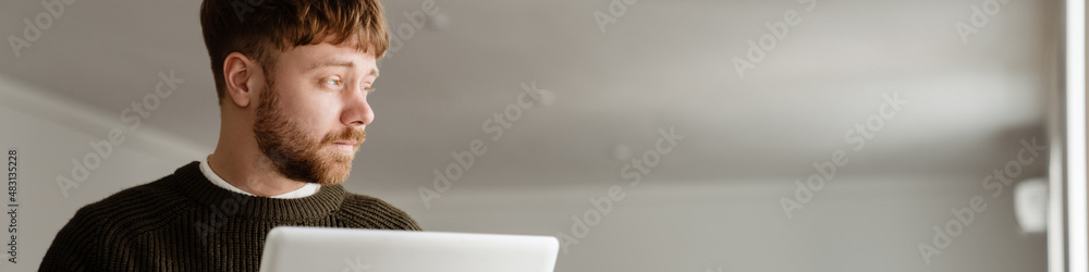 Obraz premium Young ginger man working with laptop while standing in office
