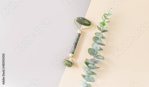 A green jade roller and a gua sha stone for facial massage and a branch of eucalyptus on a double background. Accessories for home beauty and self-care. A roller for the face. Top view, flat position.