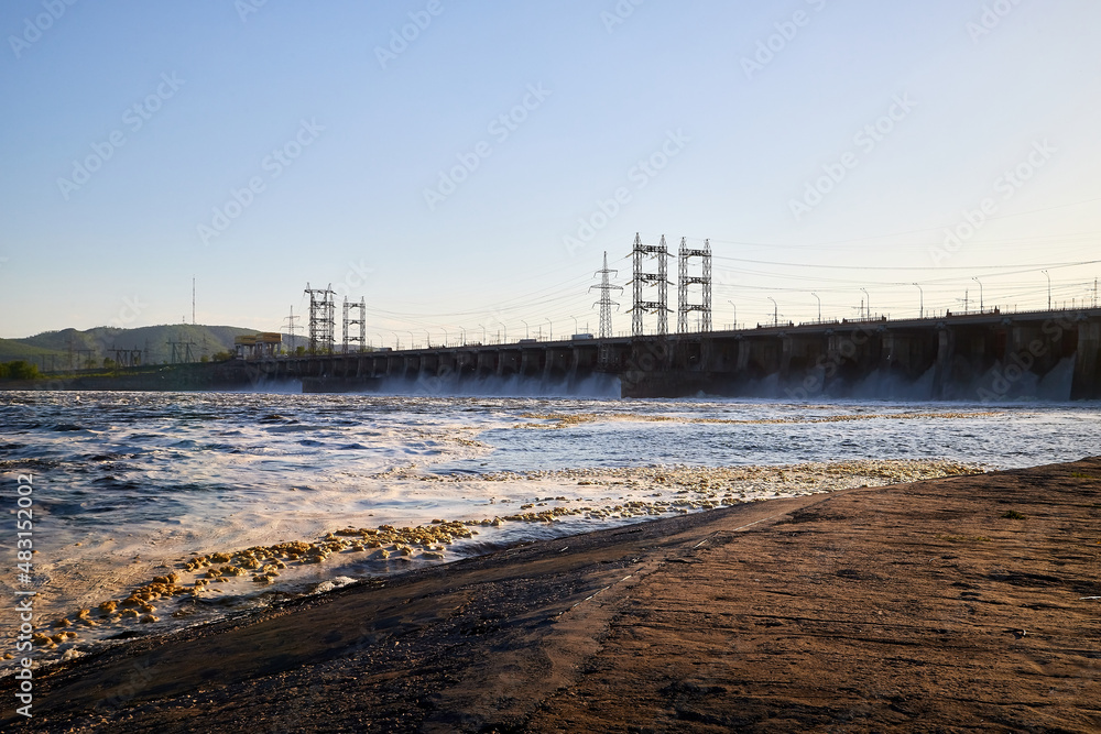 Dam of reservoir with falling water, a wave and sun on a summer or spring day. Nature landscape with beautiful water. Sea, river, lake, reservoir with blue waves during sunset. White foam on the wave