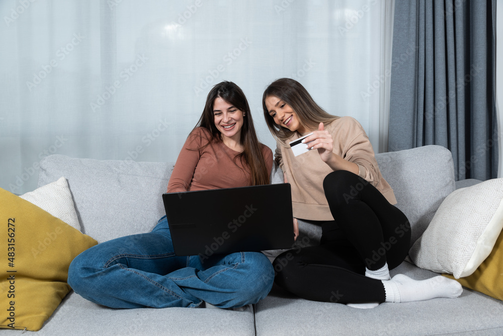 Two young happy women online buying a ticket for vacation and booking the hotel in touristic resort for holidays on laptop. Two female friends online shopping with credit card from their apartment. Two young happy women online buying a ticket for vacation and booking the hotel in touristic resort for holidays on laptop. Two female friends online shopping with credit card from their apartment.