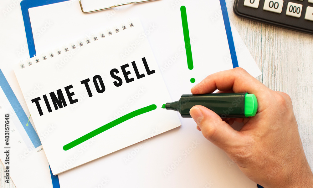 A hand with a marker writes the text TIME TO SELL in a notebook on the desktop. Top view. A hand with a marker writes the text TIME TO SELL in a notebook on the desktop. Top view.