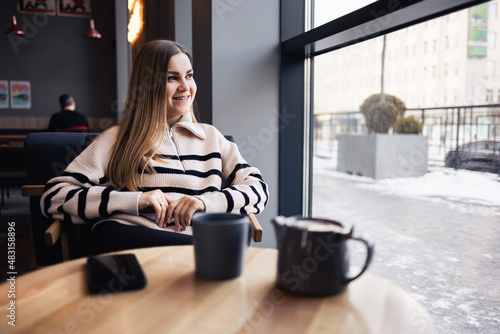 Beautiful smiling calm young woman drinking coffee looking out the window while sitting at a table in a restaurant. Morning coffee © Дмитрий Ткачук