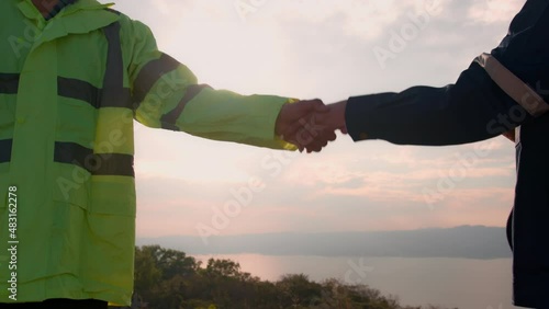 people engineer are shaking hand in a field wearing a protective helmet on her head over electrical turbines background . 