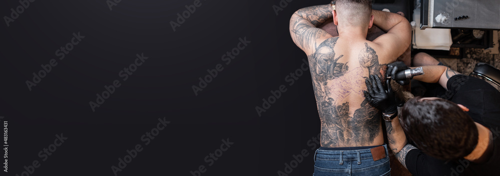 © alones - Professional tattoo artist makes a tattoo on on male back on a dark background with copy space for design and text, top view. Creative template