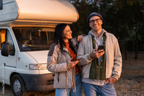 Romantic couple embracing each other while standing with smartphone and enjoying of the nature. Warm clothes on, autumn time. In background forest and car