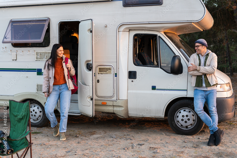 Glad loving couple is standing near the micro bus and looking at each other in the forest. Woman is looking at her man with smile Glad loving couple is standing near the micro bus and looking at each other in the forest. Woman is looking at her man with smile
