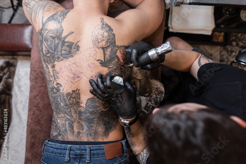 Professional male tattoo master with a tattoo machine draws a knight tattoo on a man's back, top view