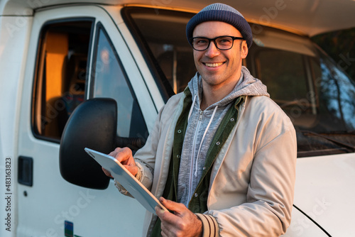 Cheerful caucasian man standing near the motor house and looking something at the tablet while spending time at the nature