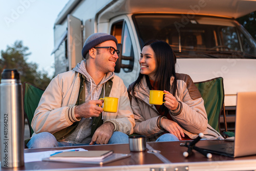 Smiling man holding cup with tea and squints against the bright sun near his woman. Happy family sitting at the nature and drinking hot tea from cups at camp fire in cozy forest