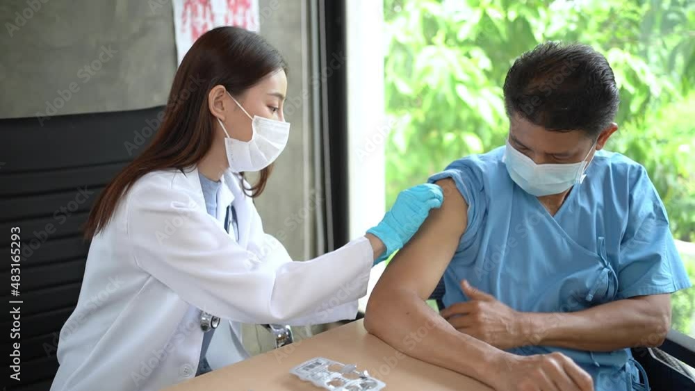 Asian female doctor in coat using cotton wiping an injection site, then vaccinated for COVID-19 and covered with bandage plaster on arm of middle-aged Asian male patient. They both wearing a face mask Asian female doctor in coat using cotton wiping an injection site, then vaccinated for COVID-19 and covered with bandage plaster on arm of middle-aged Asian male patient. They both wearing a face mask