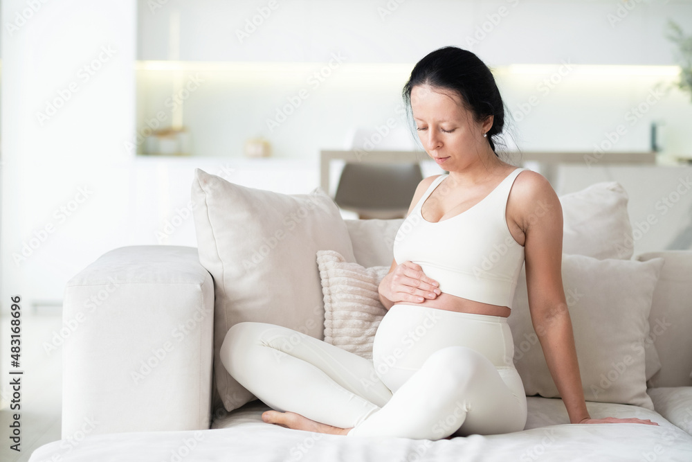 An attractive pregnant woman is sitting on a sofa in the living room of her home. She keeps her hand on her stomach so she can feel her baby move. Last months of pregnancy. An attractive pregnant woman is sitting on a sofa in the living room of her home. She keeps her hand on her stomach so she can feel her baby move. Last months of pregnancy.