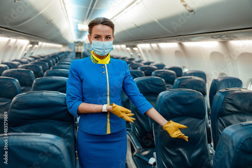 Female flight attendant wearing protective face mask and air hostess uniform while gesturing towards passenger chair in airplane