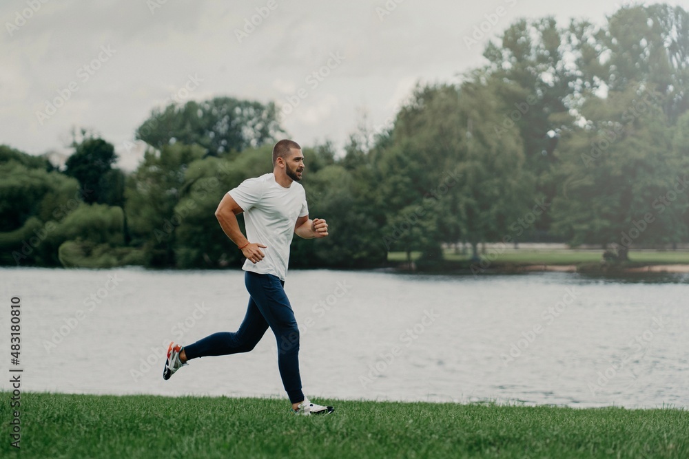 Sporty European man dressed in t shirt, trousers and sneakers and runs along river, trains outdoors, leads active lifestyle, breathes fresh air. People, sport, wellness and recreation concept. Sporty European man dressed in t shirt, trousers and sneakers and runs along river, trains outdoors, leads active lifestyle, breathes fresh air. People, sport, wellness and recreation concept.