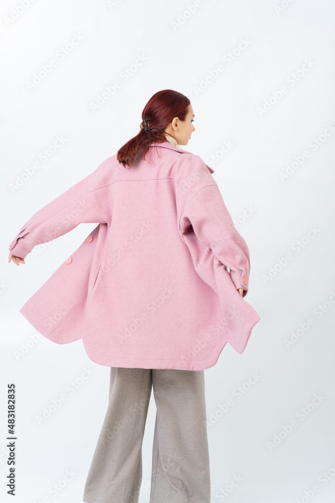 The back of a woman in a pink women coat, hands to sides, the coat flies up. Wide gray trousers made of thick fabric. Bright red hair. White background. The back of a woman in a pink women coat, hands to sides, the coat flies up. Wide gray trousers made of thick fabric. Bright red hair. White background.