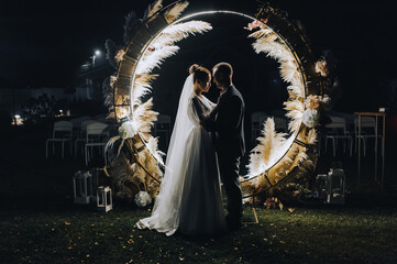 A stylish groom and a beautiful bride in a long dress are standing, hugging at night near a luminous cane arch decorated with lamps and garlands. Wedding photography, portrait.