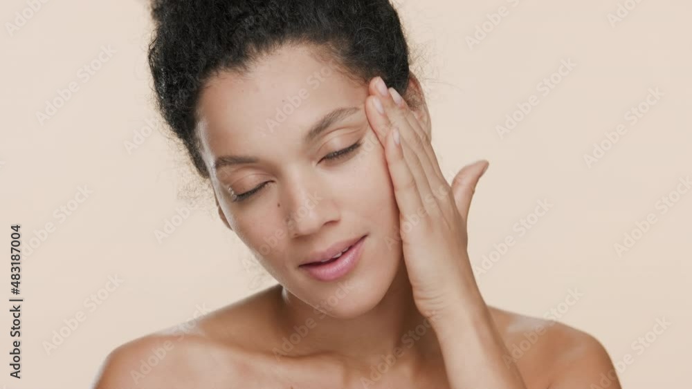 Close-up beauty portrait of good-looking young slim African American woman with black curly hair strokes her face and smiles wide for the camera on beige background | Elastic skin concept Close-up beauty portrait of good-looking young slim African American woman with black curly hair strokes her face and smiles wide for the camera on beige background | Elastic skin concept