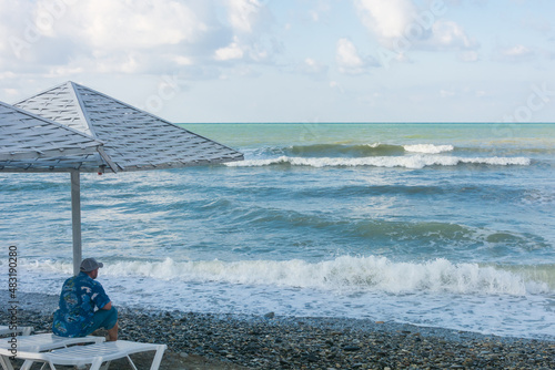 Lonely man sits on white deck chair on the beach and looks at the big waves