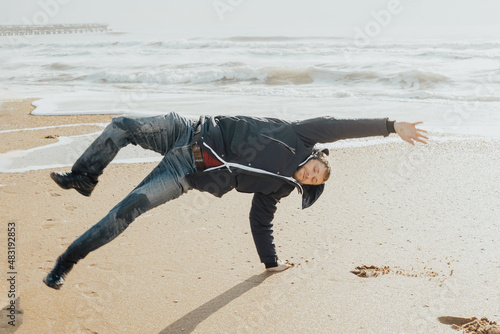 A young man in a jacket does somersaults on his hands on a sandy shore on a winter day. Travel and tourism