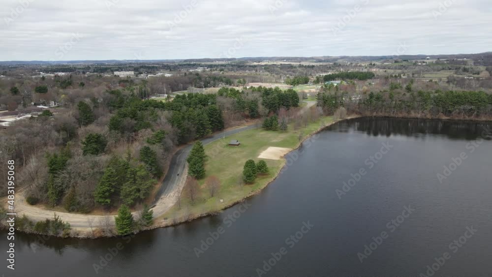 Aerial view off lake to park with woodlands, a shelter, and small empty beach. Sun reflecting off the water. Light ripples through the water. Wispy clouds. Aerial view off lake to park with woodlands, a shelter, and small empty beach. Sun reflecting off the water. Light ripples through the water. Wispy clouds.