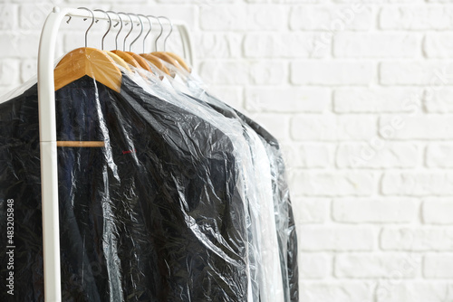 Rack with clean jackets and shirts in plastic bags on white brick background, closeup photo