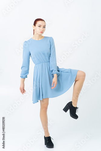 An Asian woman poses in a studio in blue summer dress and black shoes. Looks at the camera. White background.