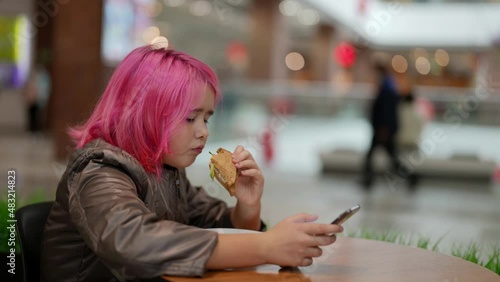 A teenage girl with pink hair is eating bread and sausage in a shopping center cafe and browsing a mobile app. A child from a dysfunctional family photo
