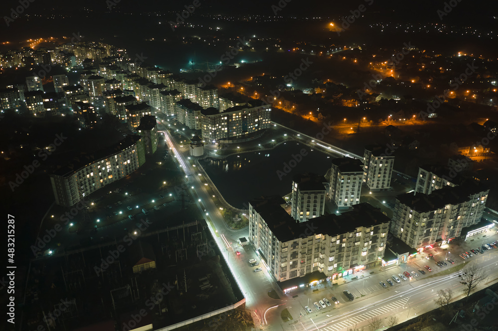 Aerial view of high rise apartment buildings and bright illuminated streets in city residential area at night. Dark urban landscape